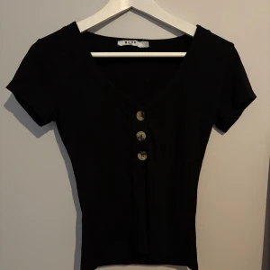 Svart ribbad topp med knappar NA-KD - Svart ribbad topp från NA-KD med v-ringning och tre snygga mönstrade knappar framtill. Kortärmad och figurnära passform, perfekt till jeans eller kjol. Materialet är mjukt och stretchigt för en bekväm känsla.
