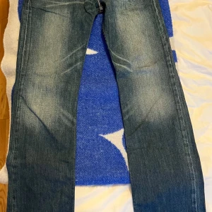 Levi's 506 Standard jeans blå - Klassiska Levi's 506 Standard jeans i blå denim med snyggt slitna detaljer och raka ben. Jeansen har fem fickor, orange sömmar och en autentisk läderpatch bak i midjan. Perfekta för en avslappnad och tidlös stil.