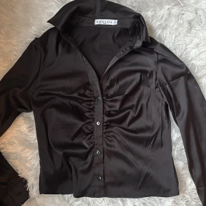 Svart satinblus från By Clara - Snygg svart blus i satin från missäy med lång ärm och knappar framtill. Blusen har en cool rynkdetalj längs knappraden och klassisk krage. Perfekt för dig som gillar en stilren men trendig look.