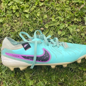 Turkosa Nike fotbollsskor med dobbar - Säljer ett par turkosa Nike fotbollsskor med svart och lila swoosh. Skorna har snörning, dobbar för gräsplan och detaljerad textur på ovandelen för bättre bollkontroll. Materialet är syntet och skon har en klassisk rund tå.