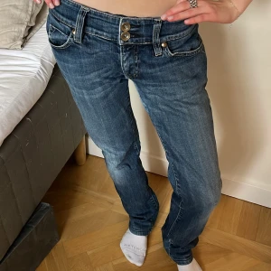 Blå Lågmidjade bootcut jeans med dubbla knappar - Snygga blå jeans med bootcut passform och låg midja. Jeansen har dubbla knappar i midjan, klassiska fickor fram och bak samt coola detaljer på bakfickorna. Tillverkade i jeansmaterial med lätt slitning för en avslappnad vibe. Dom har slitningar o skrevet, skriv privat för bilderna på slitningarna 