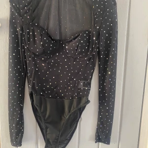 Svart glittrig body - Svart långärmad body med inbyggd korsettkänsla och glittriga paljettdetaljer över hela tyget. Toppen har markerade kupor och figurnära passform, perfekt för att sticka ut. Materialet är stretchigt och bekvämt.