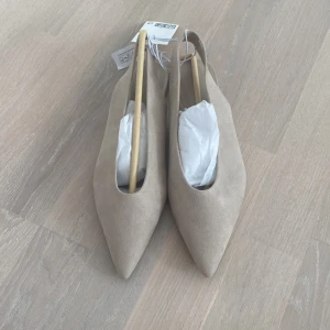 Beige slingback/ballerinaskor m. spetsig tå - Stilrena slingback/ballerinaskor i beige mocka med spetsig tå och öppen häl. Skorna har platt sula och en minimalistisk design som passar perfekt till både jeans och klänning. Snyggt och enkelt för dig som gillar en clean look.