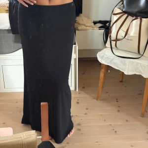 Svart långkjol i stretchigt tyg - En svart långkjol med rak passform och enkel design. Kjolen är gjord i ett stretchigt material som sitter snyggt och bekvämt på kroppen. Perfekt att styla med crop tops eller oversized tröjor för en clean look. Aldrig använd