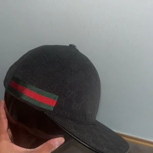 Svart Gucci keps  - Snygg svart keps från Gucci med klassiskt GG-monogram över hela kepsen. På sidan finns en ikonisk grön och röd rand. Justerbar rem baktill i svart skinn. Perfekt för dig som vill ha en exklusiv och stilren accessoar.