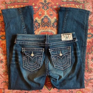 Bootcutjeans True Religion - Supersnygga jeans från True Religion. De är omsydda från skinny till bootcut, men det syns knappt (se sista bilden). De är även insydda i midjan, från storlek 28 till 27 (motsvarande S), och uppsydda i benen för att passa mig som är 160cm lång. Allt sytt professionellt av skräddare. Ena bakfickan har ett hörn som är lite uppvikt.   Jeansen är unika med ljusrosa sömmar och rosa slitningar på låren, samt knappar med rosa strass!   Mått: Innerbenslängd: 75cm Midja: 40cm Midja till gren: 20cm