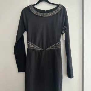 Svart klänning med pärldetaljer från Nelly - Svart kortklänning med långa ärmar och snygga silverfärgade pärldetaljer vid halsringning och midja. Klänningen har öppen rygg med coola utskärningar och dragkedja bak. Perfekt för dig som vill sticka ut med en edgy look