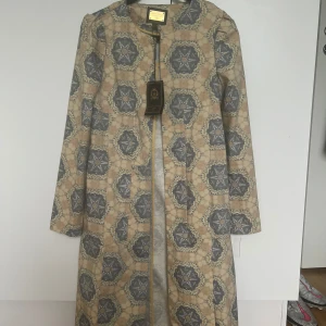 Mönstrad kappa från Ahadi Hamid - Unik kappa från Ahadi Hamid med snyggt geometriskt mönster i beige och grå toner. Långärmad och rak modell utan krage, perfekt för dig som vill sticka ut. Stängs med dold dragkedja framtill. Märke: Joyeux Noël, Tehran.