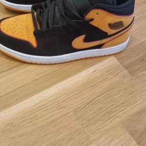 Nike Air Jordan 1 svart/gul sneakers - Snygga Nike Air Jordan 1 sneakers i svart och gul med klassisk siluett. Skorna har svarta snören, vit mellansula och gul detalj på tån, sidorna och hälen. Perfekta för dig som vill sticka ut med en ikonisk streetstyle-look.