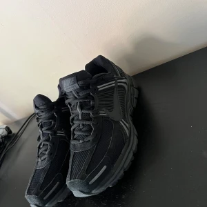 Svarta Nike vomero zoom 5 sneakers med mesh och detaljer - Snygga helsvarta Nike vomero zoom 5 sneakers med meshpaneler och coola detaljer i syntet. Skorna har en chunky sula, snörning och diskreta reflexdetaljer. Perfekta för dig som gillar sportig och modern stil. Swoosh-logga på sidan och mönstrad häl.