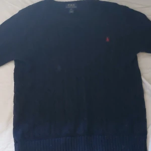 Mörkblå stickad tröja Polo Ralph Lauren - Snygg mörkblå stickad tröja från Polo Ralph Lauren med klassisk kabelstickning och röd broderad logga på bröstet. Tröjan har rund halsringning och långa ärmar, perfekt för lager-på-lager-stil.