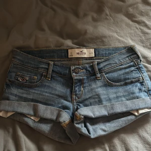 Blå jeansshorts från Hollister - Snygga blå jeansshorts från Hollister med klassisk femficksdesign, uppvikta benslut och slitna detaljer. Shortsen har låg midja och är tillverkade i mjukt denim med kontrastsömmar och broderad logga på fickan. Perfekta för varma dagar och en avslappnad stil. Storlek w24/XXS