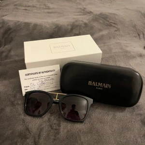 Svarta solglasögon från Balmain - Snygga svarta solglasögon från Balmain med fyrkantig form och cool gulddetalj över näsryggen. Glasen är mörka och bågarna är robusta med en exklusiv känsla. Kommer med originaletui och äkthetsintyg.