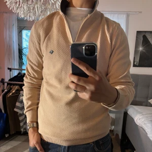 Beige half zip tröja Sir Raymond Tailor - Snygg beige långärmad tröja från Sir Raymond Tailor med half zip och mörk ribbad krage. Tröjan har diskret strukturmönster och liten logga på bröstet. Perfekt till jeans för en clean och stilren look.