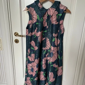 Blommig ärmlös klänning med krage - Snygg ärmlös klänning med stor krage och blommigt mönster i rosa, grönt och blått på mörk botten. Klänningen har en rak, något A-linjeformad siluett och dragkedja bak. Perfekt för dig som gillar färg och retrovibbar!