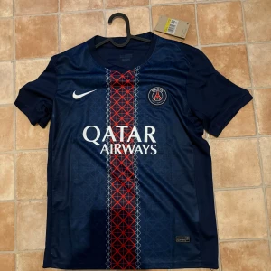 PSG Vitinha 17 matchtröja Nike - Paris Saint-Germain fotbollströja med Vitinha 17-tryck, officiell Champions League badge på ärmen och Qatar Airways sponsor. Djupblå med röd och vit rand i mitten, PSG-emblem på bröstet och Nike-logga. Tillverkad i lätt och ventilerande material, perfekt för fotbollsälskare.