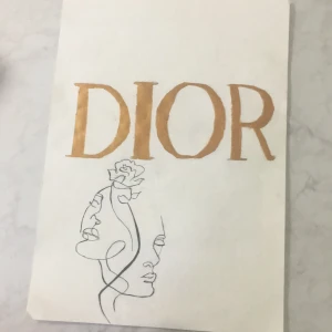 DIOR målning  - Stilren bok med handritad linjekonst av två ansikten och en blomma, samt den ikoniska texten 'DIOR' i guld. Perfekt för dig som älskar mode, konst och unika illustrationer. Passar som inspiration eller coffee table-bok för unga kreativa.
