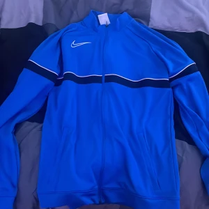 Blå Nike Dri-Fit vindjacka XL - Snygg blå Nike Dri-Fit vindjacka med hel dragkedja och vit broderad swoosh på bröstet. Jackan har svarta och vita detaljer över bröstet och är tillverkad i ett lätt, sportigt material. Perfekt för dig som gillar sportig stil och vill ha något bekvämt och stilrent.