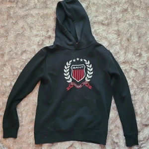 Svart hoodie från GANT med emblem - Svart hoodie från GANT med stort broderat emblem i vitt och rött på bröstet. Klassisk huva, känguruficka och ribbade muddar. Tillverkad i mjuk bomullsblandning med polyester. Perfekt för dig som gillar stilren streetwear med preppy touch.