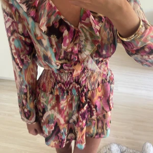 Färgglad kortklänning med volang - Superfin kortklänning med färgstarkt blommigt mönster i rosa, lila, turkos och beige toner. Klänningen har långärmade transparenta ärmar, v-ringning med volangdetalj och smockad midja. 