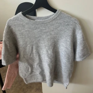 Grå stickad tröja från Gina Tricot - Supermjuk grå stickad tröja från Gina Tricot med korta ärmar och rund halsringning. Perfekt för lager-på-lager och chill vardagsstil. Enkel design som funkar till allt och är lätt att matcha med dina favoritjeans eller kjol.