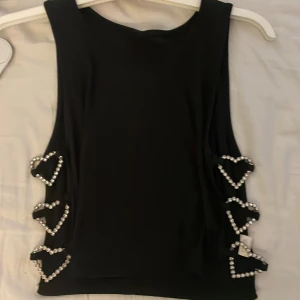 Svart ärmlös topp med hjärt-detaljer - Supersnygg svart ärmlös topp med öppna sidor där tre hjärtformade detaljer i silverfärgade stenar sitter på varje sida. Toppens tyg är lätt ribbat och ger en cool look. Perfekt för dig som vill sticka ut med glittriga detaljer och unik design.