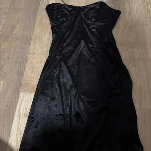 Svart långklänning från Divided - Supersnygg svart långklänning från Divided med glansigt tyg och smala axelband i kedjedesign. Klänningen har en figurnära passform upptill och smock i ryggen för extra komfort. 