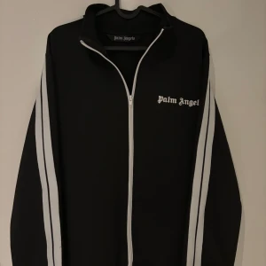 Palm Angels svart vindjacka med ränder - Svart vindjacka från Palm Angels med vita ränder längs ärmarna och vit dragkedja. Jackan har hög krage och logga på bröstet. Perfekt för en sportig och trendig look.
