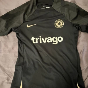Chelsea träningströja - Chelsea FC svart fotbollströja från Nike i slim fit-modell. Tröjan har korta ärmar, Dri-Fit-material som andas och tryckt trivago-logga på bröstet. Chelsea-emblem och Nike swoosh i vitt framtill. Perfekt för träning eller match. Har blivit för liten för mig. STORLEK L I BARN, inte Herr