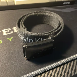 Svart tygbälte Calvin Klein Jeans - Svart tygbälte från Calvin Klein Jeans med tydlig logga längs bandet och en svart metallspänne. Bältet har en clean och modern look, perfekt för att lyfta jeans eller casual outfits. Justerbar längd och klassisk design med rektangulärt spänne.