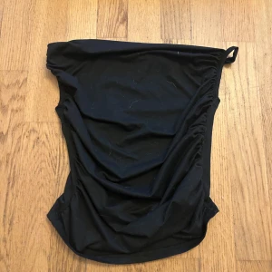 Svart bandeau tubtopp - Säljer en svart bandeau tubtopp med draperad effekt på ena sidan och en liten ögla upptill. Toppen är axelbandslös och har en tight passform, perfekt för att skapa en clean och stilren look.