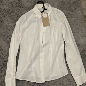 Vit klassisk skjorta från Soyaconcept - Snygg vit skjorta från Soyaconcept med klassisk krage och knäppning framtill. Skjortan har lång ärm och figurnära passform, perfekt till jeans eller kostymbyxor. Tillverkad i återvunnen polyester för en modern känsla.