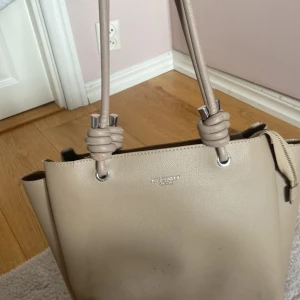 Beige axelväska från Art Street london - Stilren beige axelväska med snygga knutdetaljer på handtagen och silvriga metalldelar. Väskan har flera fack, dragkedja i mitten och är tillverkad i slätt syntetmaterial. Perfekt för dig som vill ha en praktisk och trendig väska med mycket plats.
