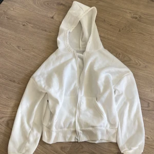 Vit hoodie med dragkedja och huva - Snygg vit hoodie med dragkedja framtill och stor huva. Tröjan har långa ärmar, ribbade muddar och fickor på sidorna. Perfekt för en chill och avslappnad stil. Passar till allt och är superenkel att matcha med andra plagg.