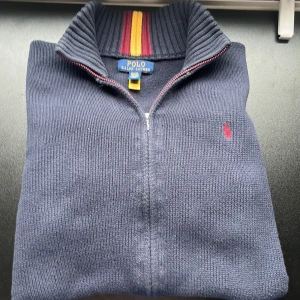 Mörkblå stickad tröja Polo Ralph Lauren - Mörkblå stickad kofta från Polo Ralph Lauren med hel zip och ribbad krage med röda och gula ränder. Broderad röd logga på bröstet och breda muddar vid ärmslut och nederkant. Perfekt för dig som gillar klassisk och sportig stil.