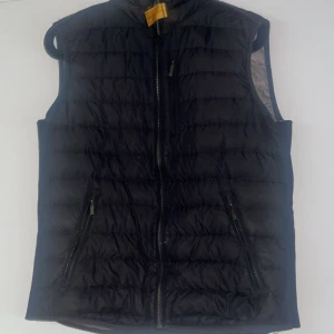 Svart dunväst från Parajumpers - Säljer en svart dunväst från Parajumpers med quiltad design och dragkedja framtill. Västen har två fickor med dragkedja och en hög krage. Superlätt och perfekt för lager-på-lager-stil. Märkeslogga på nacken och insidan. På baksidan har den gått lite sönder men jag syde ihop den och den sitter fast jättebra och man märker knappast av den.