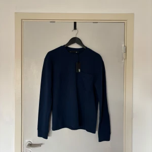 Mörkblå sweatshirt från 157 - En stilren mörkblå sweatshirt från 157 med rund halsringning och en bröstficka. Tröjan är i mjuk bomull och har långa ärmar med ribbade muddar. Perfekt för dig som gillar enkel och clean stil.