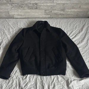 Svart bomberjacka från Zara, storlek L - Stilren svart bomberjacka från Zara i storlek L. Jackan har klassisk krage, knäppning med knappar framtill och två snedställda fickor. Ribbstickade muddar vid ärmslut och nederkant ger en snygg passform. Perfekt för dig som gillar en clean och tidlös look.