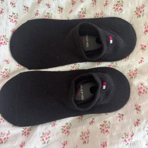 Svarta ankelstrumpor Tommy Hilfiger - Svarta ankelstrumpor från Tommy Hilfiger med diskret logga i rött, vitt och blått vid öppningen. Strumporna är låga och har anti-slip häl för att sitta på plats. Ett par för 30kr, två för 50kr