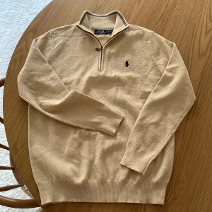 Beige half zip tröja från Polo Ralph Lauren - Snygg beige tröja från Polo Ralph Lauren med half zip och klassisk krage. Tröjan har lång ärm, diskret struktur i materialet och den ikoniska broderade loggan på bröstet. Perfekt för dig som gillar stilren och tidlös design. Jag är själv M och den passar men passar nog precis L så de beror på hur man vill att den ska sitta, därav M/L storlek.