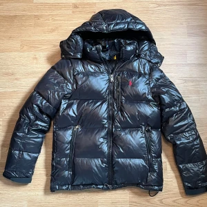 Svart dunjacka Polo Ralph Lauren - Snygg svart dunjacka från Polo Ralph Lauren med avtagbar huva och glansig finish. Jackan har dragkedjor på fickorna, en röd logga på bröstet och quiltad design. Perfekt för kalla dagar när du vill hålla stilen och värmen.