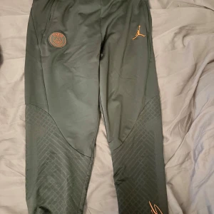 Jordan psg byxor - Mörkgröna träningsbyxor från Jordan i samarbete med PSG. Byxorna har orangea loggor, smal passform och snygga detaljer på benen. Tillverkade i polyester, perfekta för sport eller chill. Elastisk midja och diskret mönster på nedre delen av benen. Har blivit like för små för mig. Köpta från nike.com