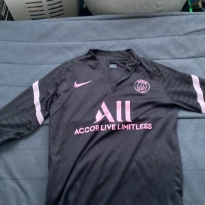 PSG långärmad svart Nike Dri-Fit tröja - Säljer en svart långärmad Paris Saint-Germain fotbollströja från Nike med rosa detaljer och klubbmärke på bröstet. Tröjan har Dri-Fit-material och subtila zebramönster på ärmarna. Snygg V-ringning och tryck med 'Accor Live Limitless' på framsidan. Kedjan är sönder dock men allt annat e perfekt