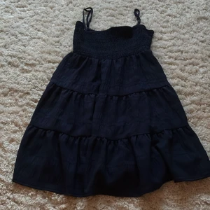 Marinblå volangklänning - Supersöt marinblå kortklänning med smockad överdel och tunna axelband. Kjolen har flera lager volanger som ger en flowy känsla. Perfekt för varma dagar när du vill känna dig både chill och snygg.