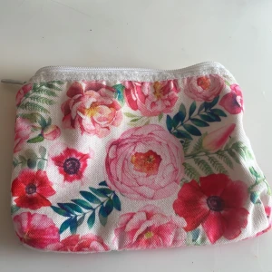 Blommig liten necessär med dragkedja - Söt liten necessär med blommigt mönster i rosa, rött och grönt på vit botten. Perfekt för smink eller småprylar. Necessär har dragkedja upptill och är gjord i ett tygmaterial med ljus insida.