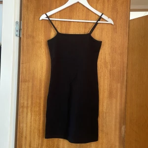Svart tight kort klänning med smala axelband - Enkel och stilren svart kort klänning med smala axelband och rak halsringning.
