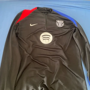 FC Barcelona långärmad träningströja Nike - Snygg svart långärmad träningströja från Nike med FC Barcelona-logga och Spotify-sponsor på bröstet. Tröjan har dragkedja vid halsen och detaljer i rött och blått på axlarna. Tillverkad i ett lätt och sportigt material som andas.