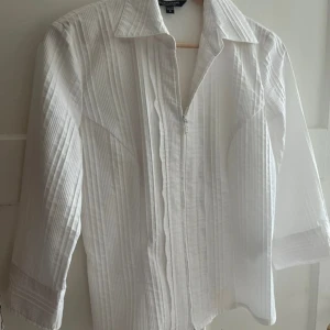 Vit skjorta med dragkedja från Brandtex - Vit skjorta från Brandtex med diskret randigt mönster i strukturen. Skjortan har krage, trekvartsärm och stängs med dragkedja framtill. Snygg och stilren modell som passar till många olika looks.