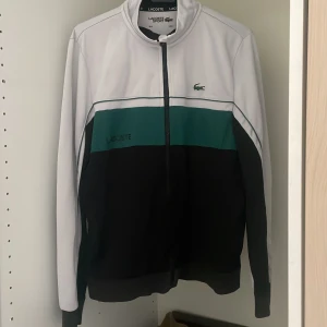 Lacoste zip  - Snygg vindjacka från Lacoste Sport i färgblock med vitt upptill, grönt i mitten och svart nedtill. Klassisk ståkrage, hel dragkedja och broderad Lacoste-logga på bröstet. Perfekt för en sportig och clean look.