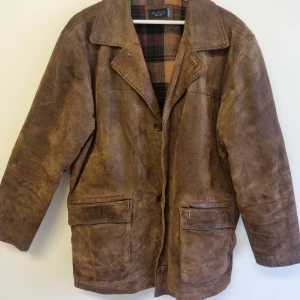 Brun mockajacka från Wilhelm - Snygg brun mockajacka från Wilhelm med klassisk krage och stora fickor med dragkedja. Jackan har rutigt innerfoder i bruna och svarta toner, perfekt för dig som gillar vintagevibbar och tidlös stil. Passar dig som vill ha en jacka med lite längre modell och avslappnad känsla.